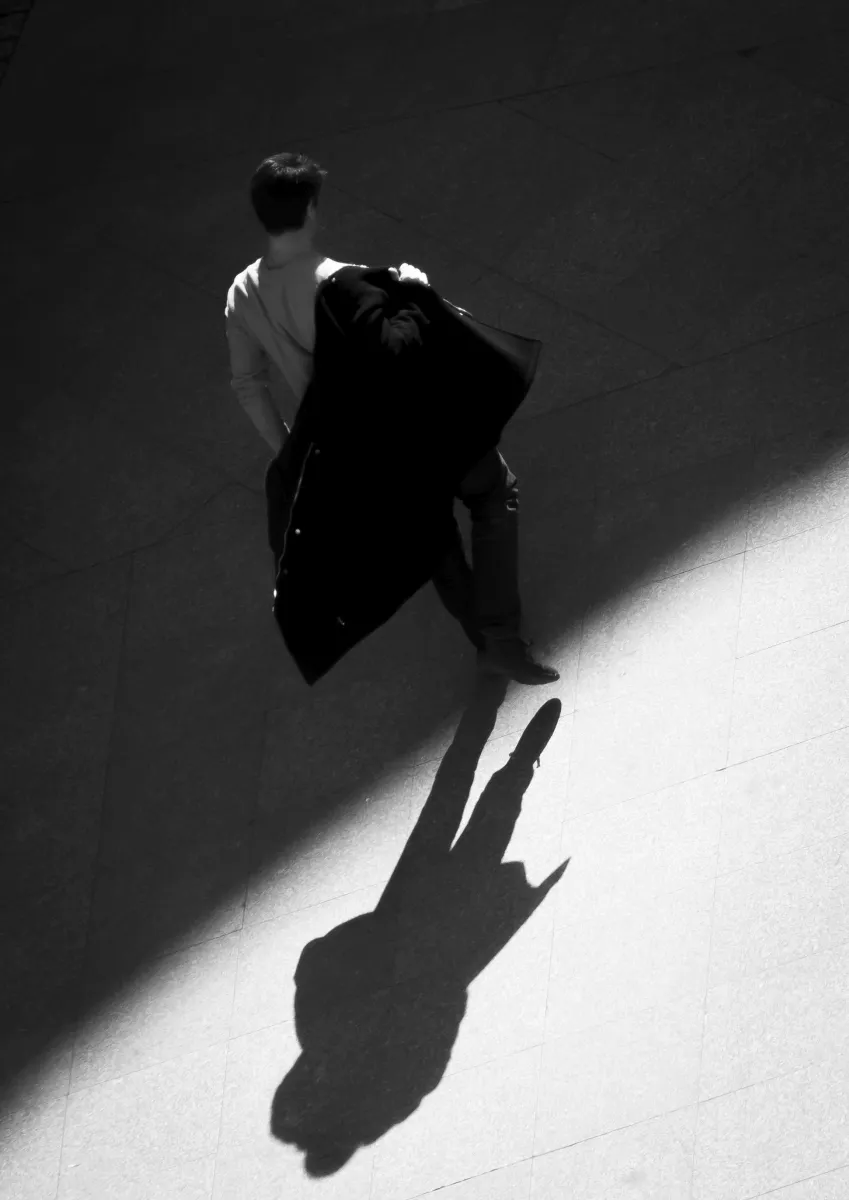 Person carrying a dark coat and casting a long shadow across sunlit pavement